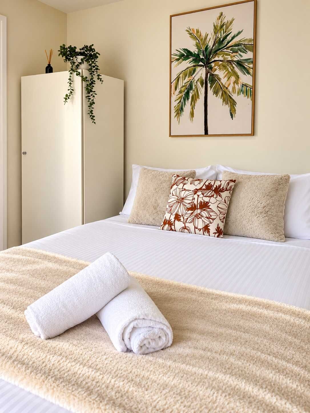Een netjes opgemaakt bed met een opgerolde witte handdoek, een bloemenkussen en een ingelijst foto van een palmboom aan de muur.