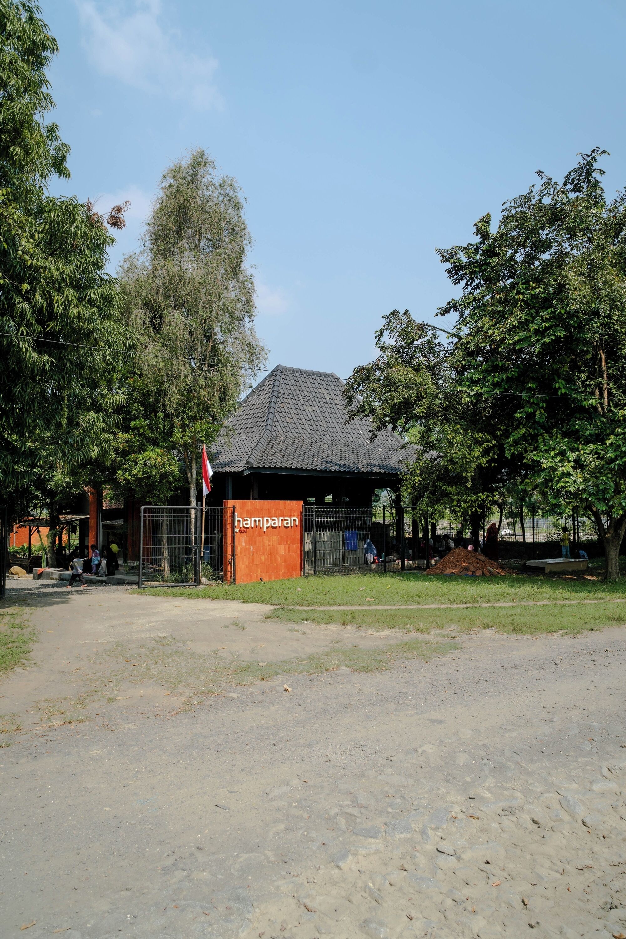 Een gebouw met een bord waarop 'hamparan' staat, voor een omheind gebied met bomen.