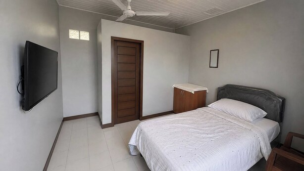 A bedroom with a bed, a television mounted on the wall, a ceiling fan, a wooden door, and a small wooden cabinet. - Nata Hotel Tasikmalaya (Tasikmalaya)