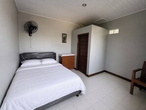 Une chambre à coucher avec un lit, une armoire en bois, une porte, un ventilateur et un petit tableau accroché au mur.