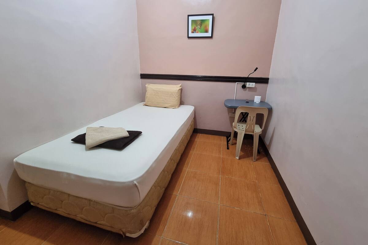 A small, neatly arranged room with a single bed, a chair, a small table, and a wall-mounted power outlet.