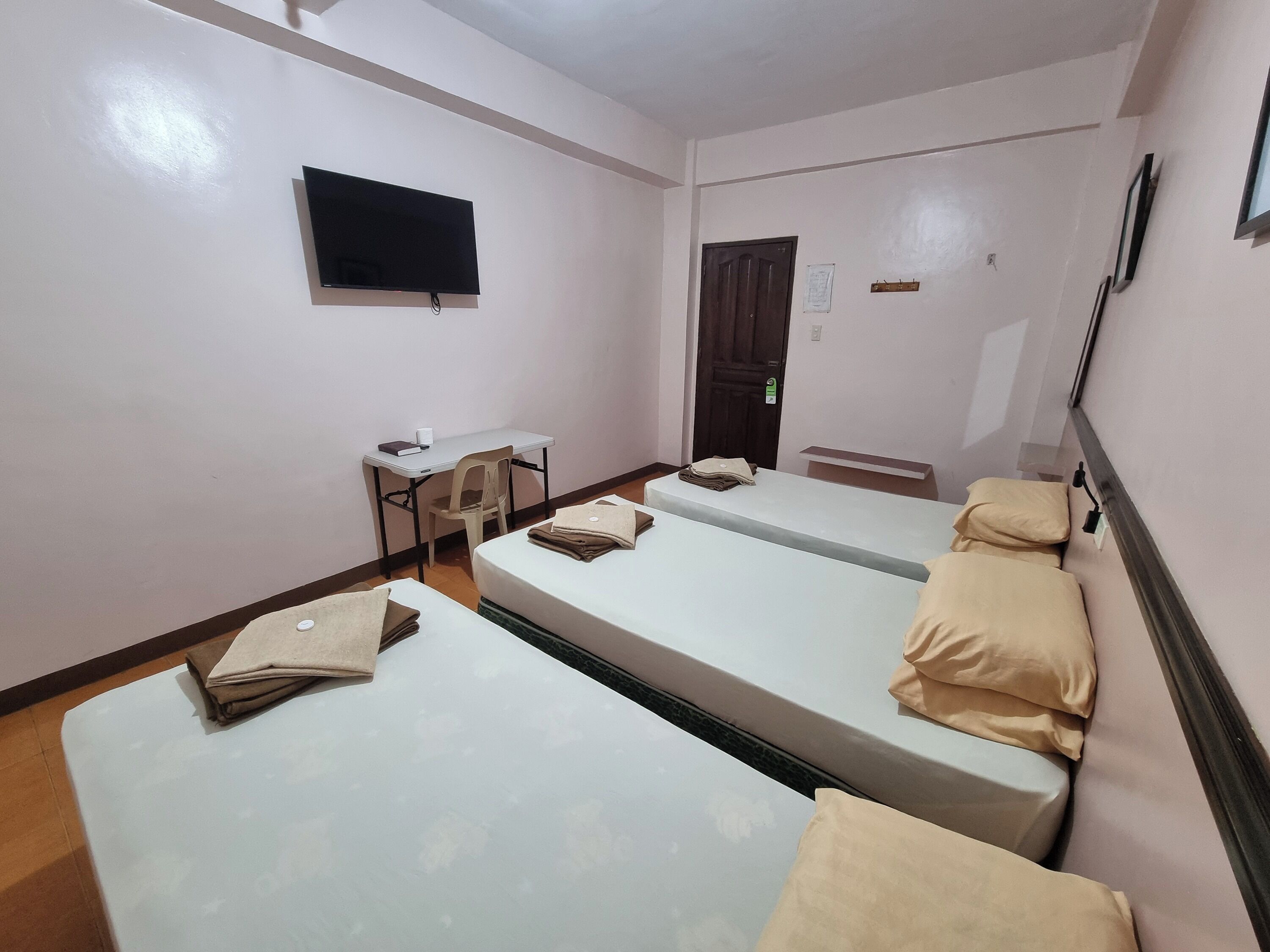 A hotel room with three single beds, a desk with a chair, a flat-screen TV, and a door.