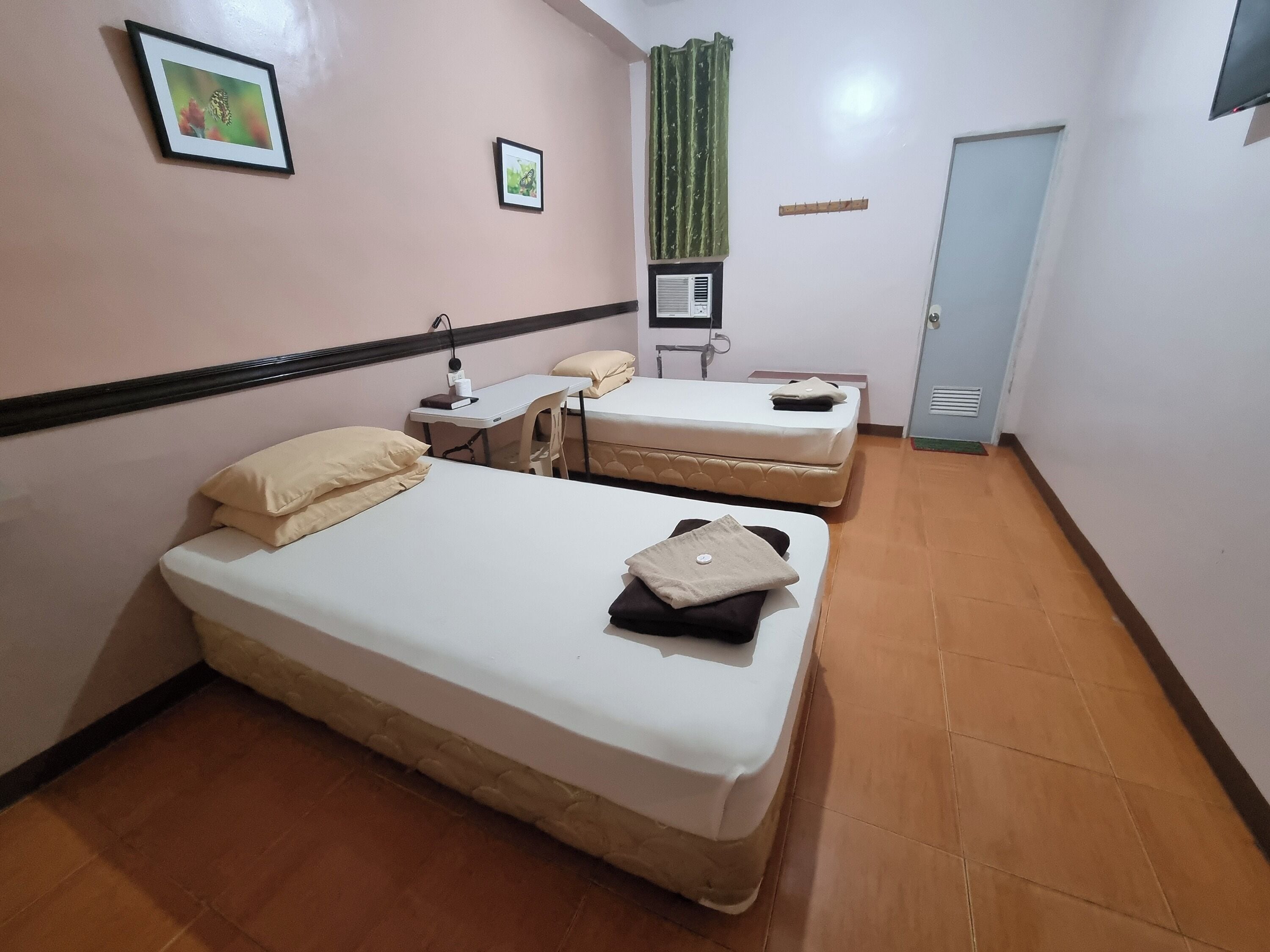 Habitació d'hotel amb dos llits, un escriptori, una cadira, una finestra amb cortines, una porta i un aire condicionat fixat a la paret.