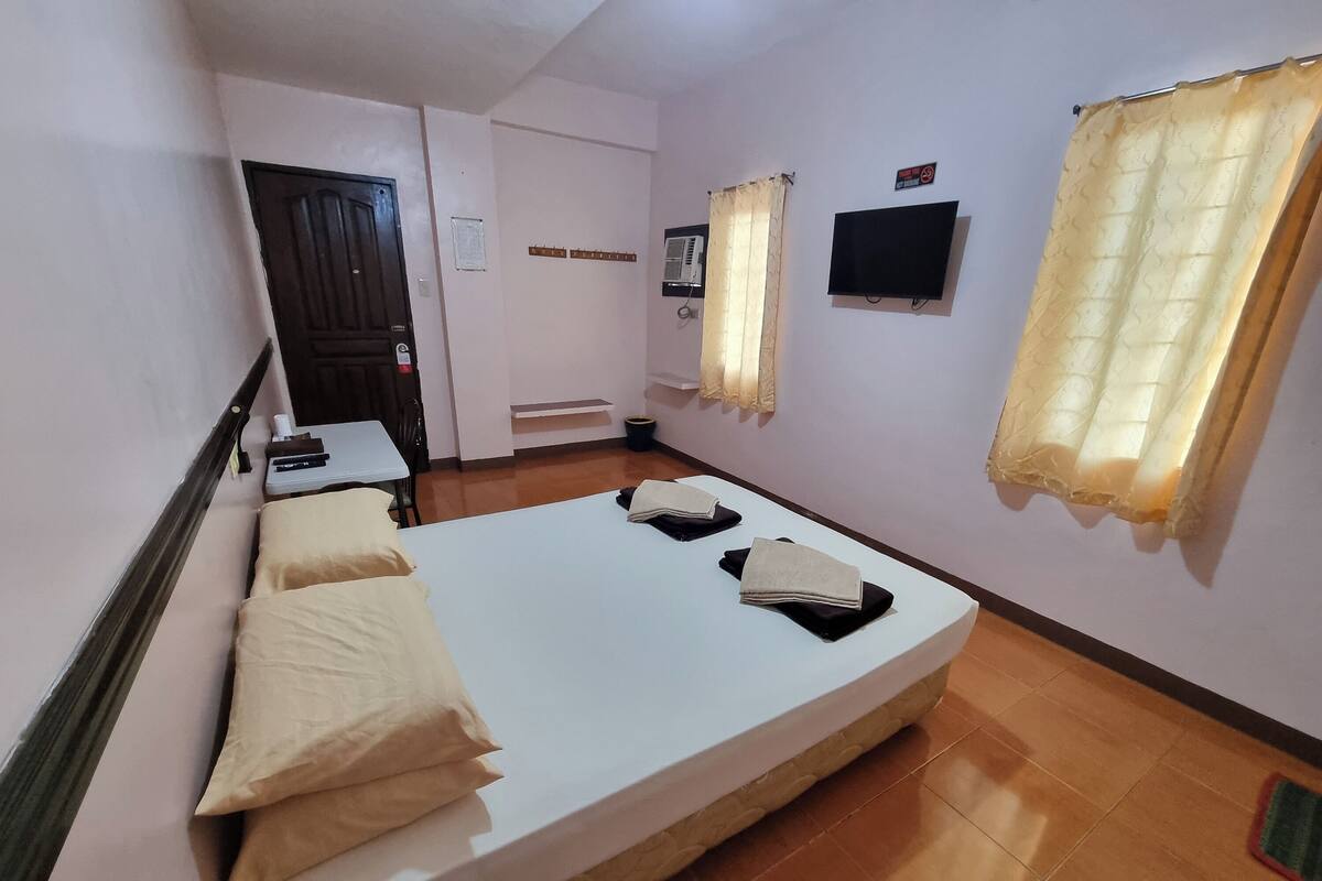 A room with a bed, a desk, a chair, a television, and a window with curtains.