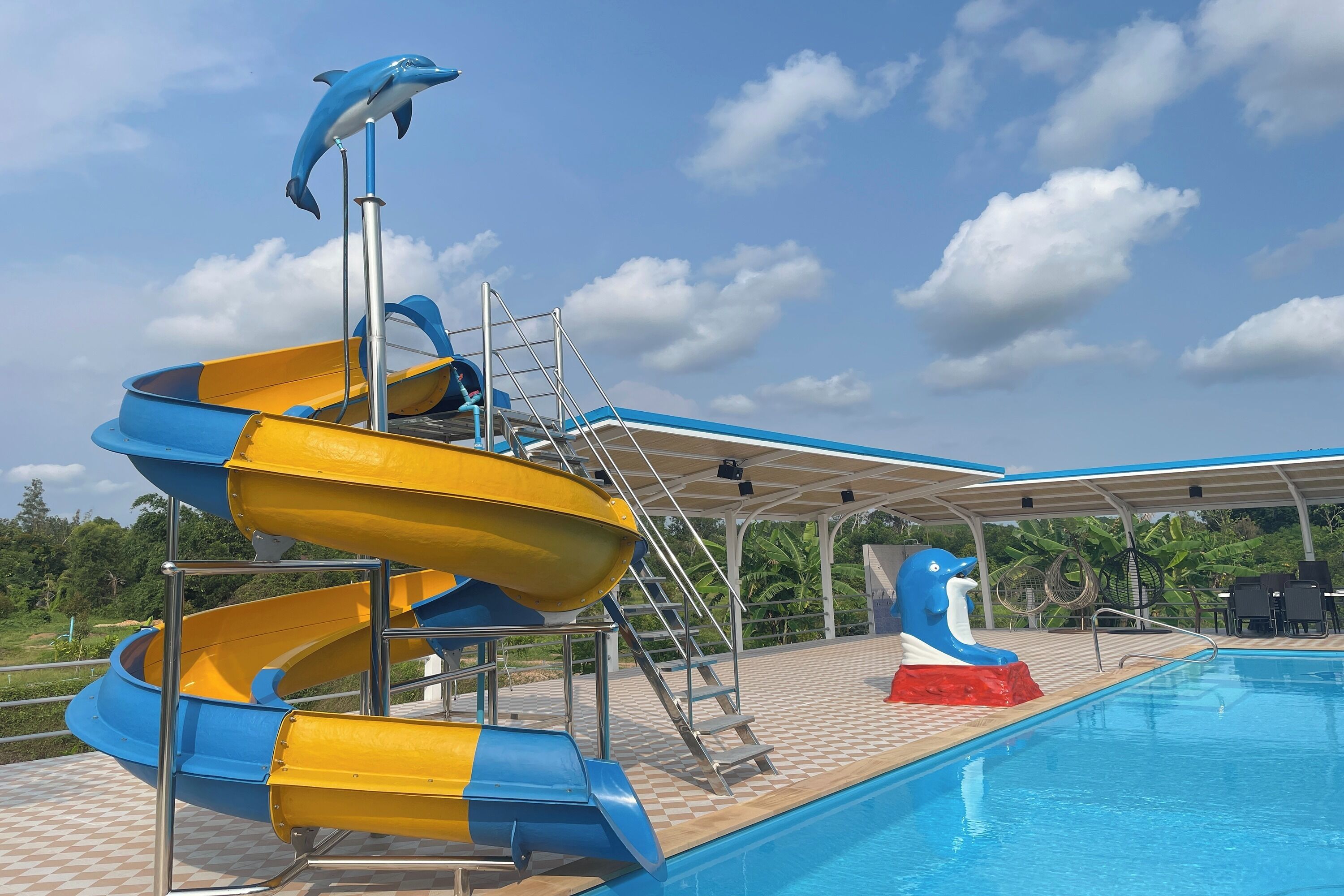 A water slide with a dolphin figure at the top, a pool, and a covered area with chairs and tables.