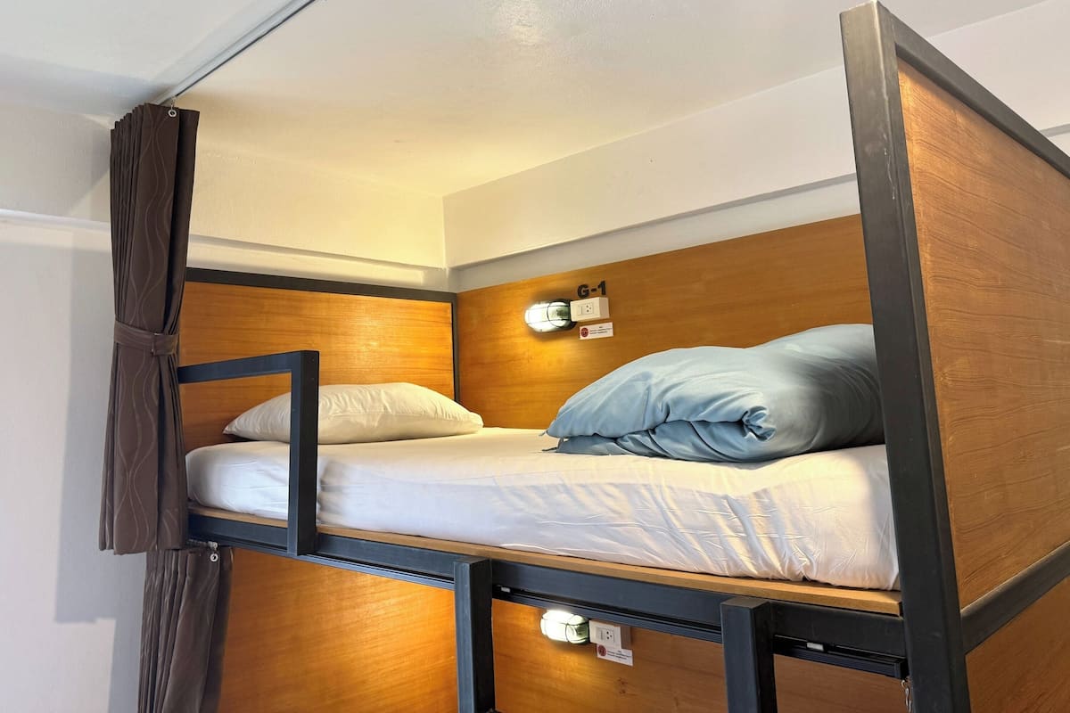 Uma cama de solteiro com peseira de madeira, duas almofadas, uma luminária fixada na parede e uma tomada.