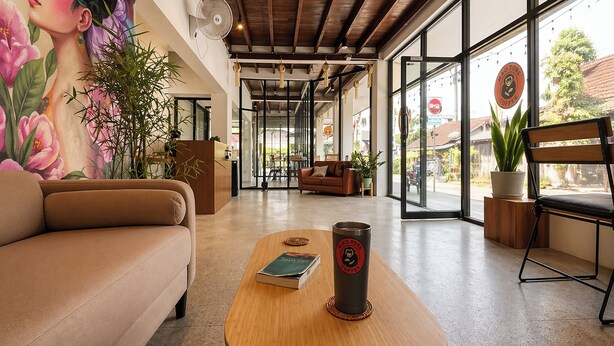 A modern interior with a sofa, a wooden table with a book and a coffee cup, a mural, and large windows. - Lazy Gibbon 2 Chiang Mai - Haiya (Chiang Mai)