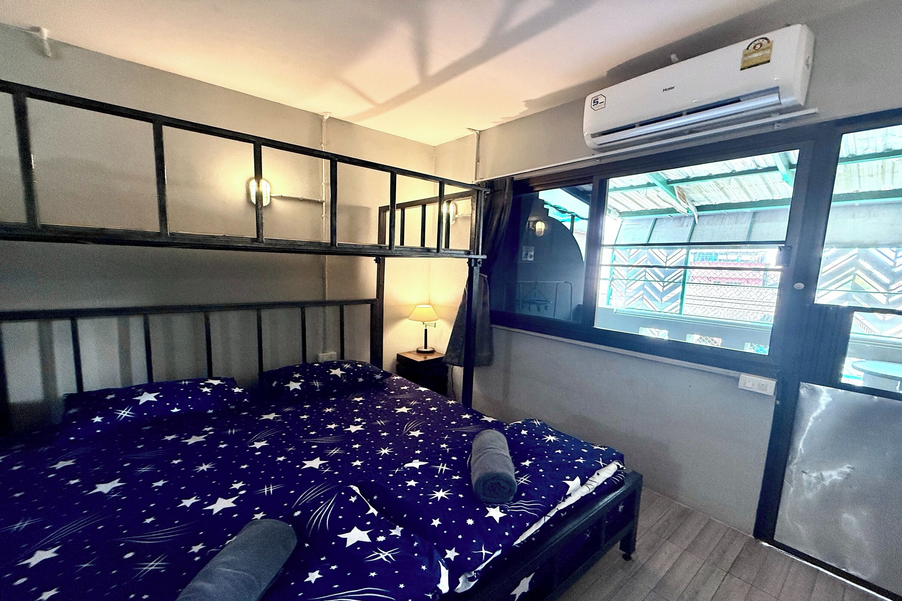 Una habitación con cama, ventana, aire acondicionado y mesita de noche con lámpara.