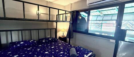 Una habitación con cama, ventana, aire acondicionado y mesita de noche con lámpara.