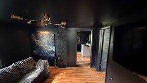 A modern interior with a wooden floor, a sofa, a painting, and a kitchen in the background. - Dark & Cozy Vibez with Appalachian Mountain Views (Candler)