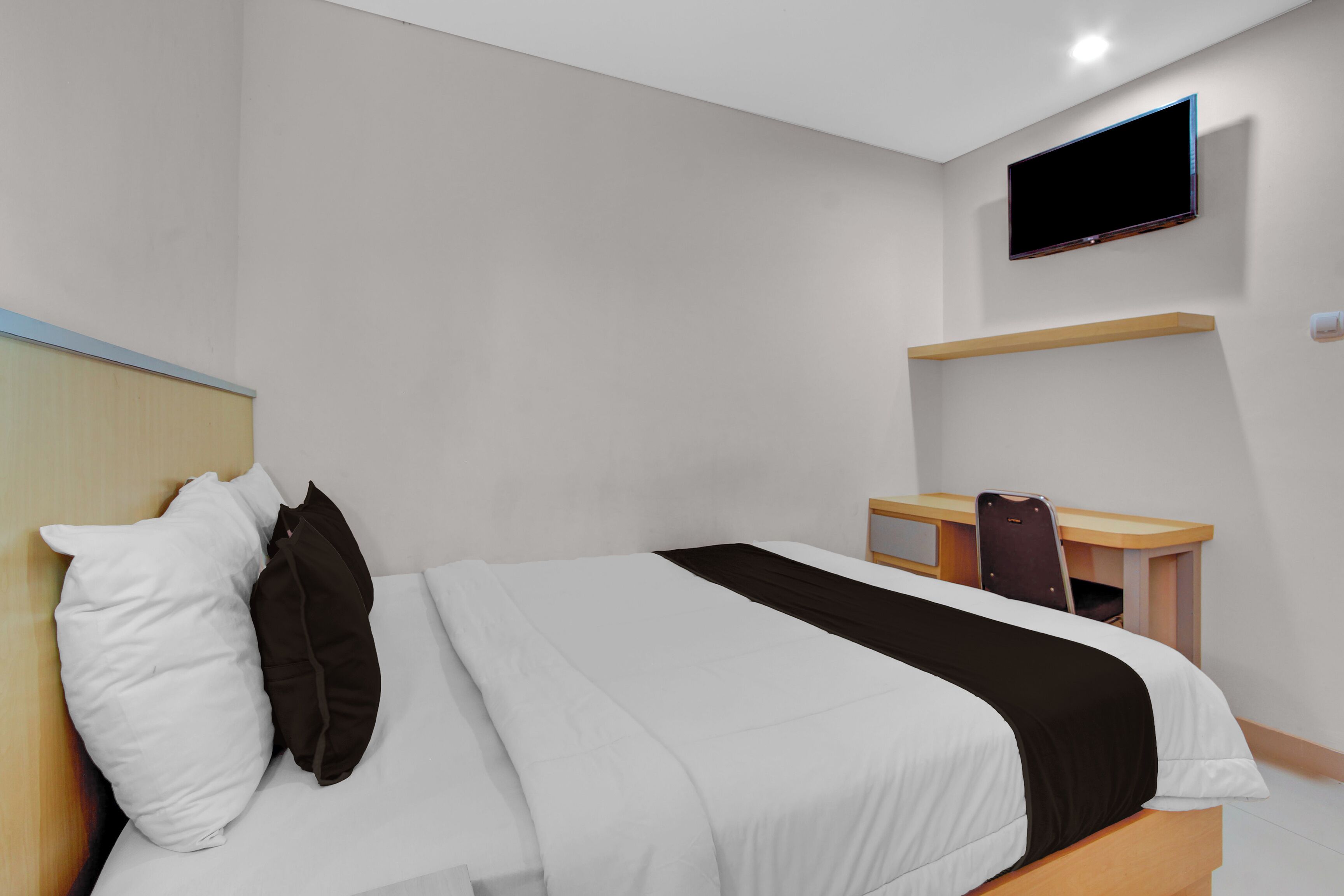 A hotel room with a bed, a desk, a chair, a TV mounted on the wall, and a shelf.