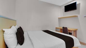 A hotel room with a bed, a desk, a chair, a TV mounted on the wall, and a shelf.