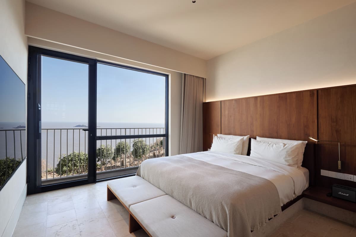 Un dormitorio moderno con una cama grande, una banca, una mesita de noche y vista al mar desde un balcón.