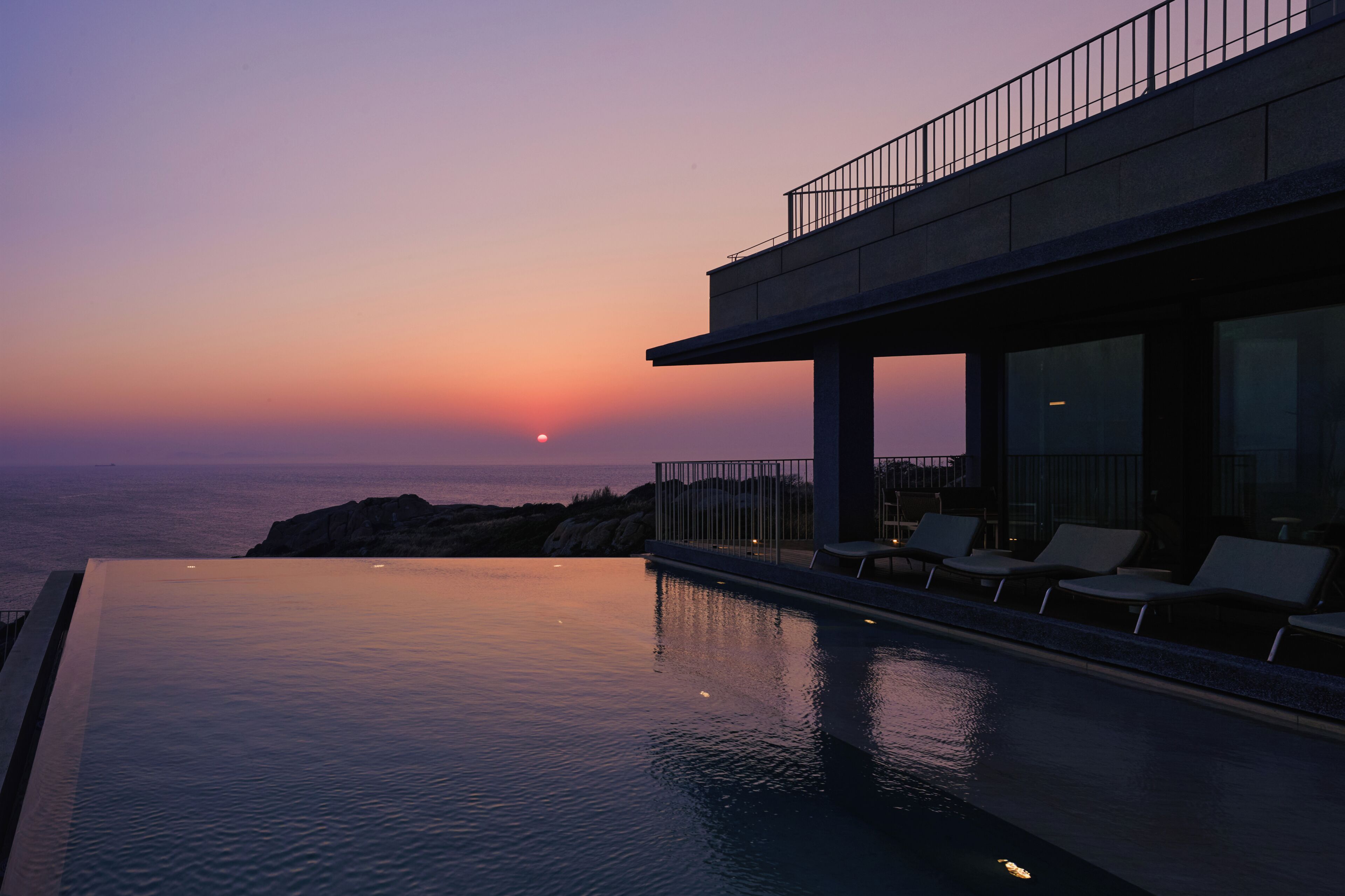 Une piscine à débordement moderne offrant une vue sur l’océan au coucher du soleil, un balcon avec des sièges et un ciel dégagé.