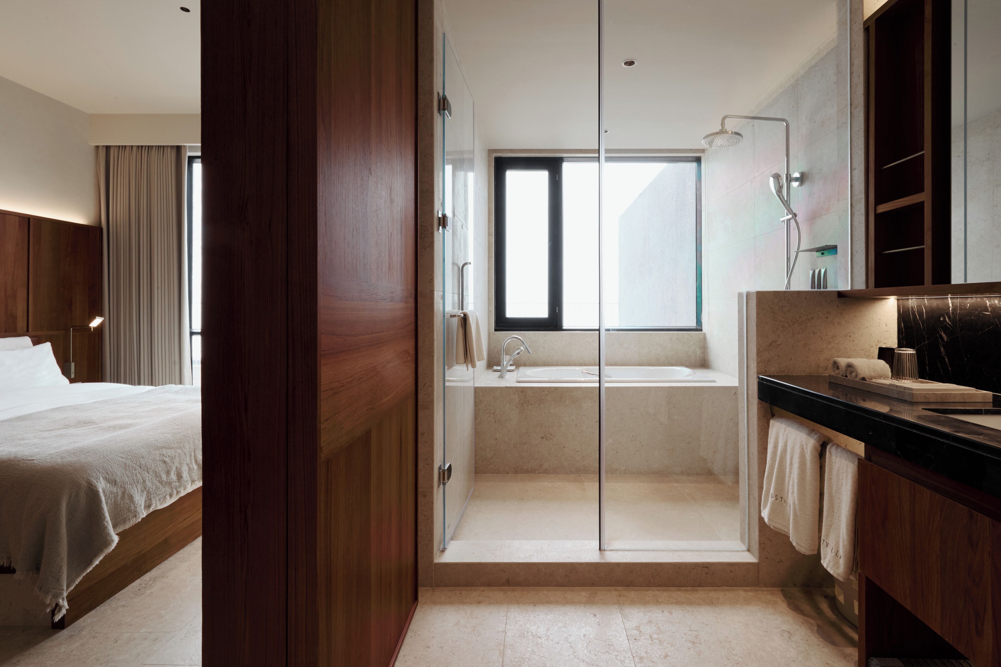 A modern hotel room with a wooden bed, a glass-enclosed shower, a bathtub, and a large window providing natural light.