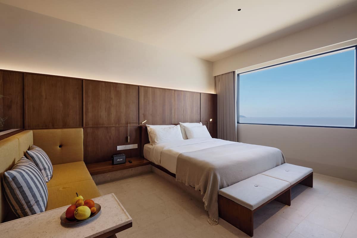 Una habitación de hotel moderna con cama, sofá, una mesita con fruta y un ventanal grande.