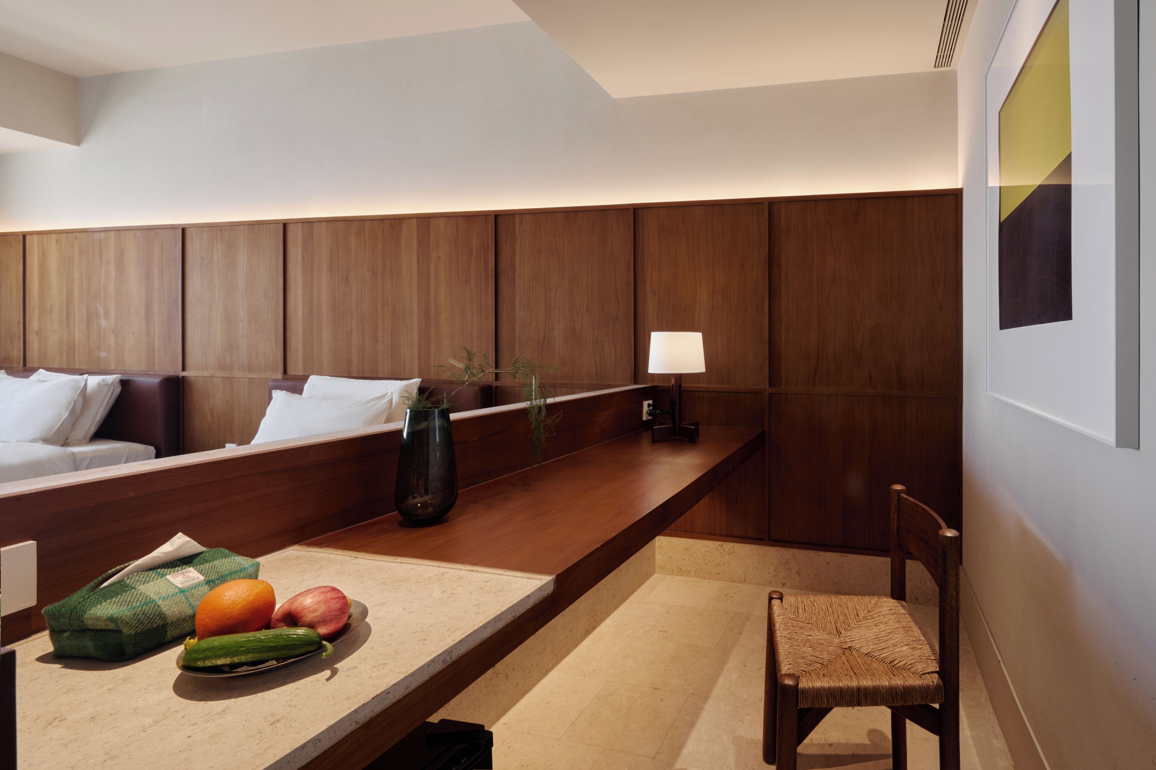 A modern hotel room with a wooden headboard, a bedside table with fruits and a vase, a chair, and a wall-mounted lamp.