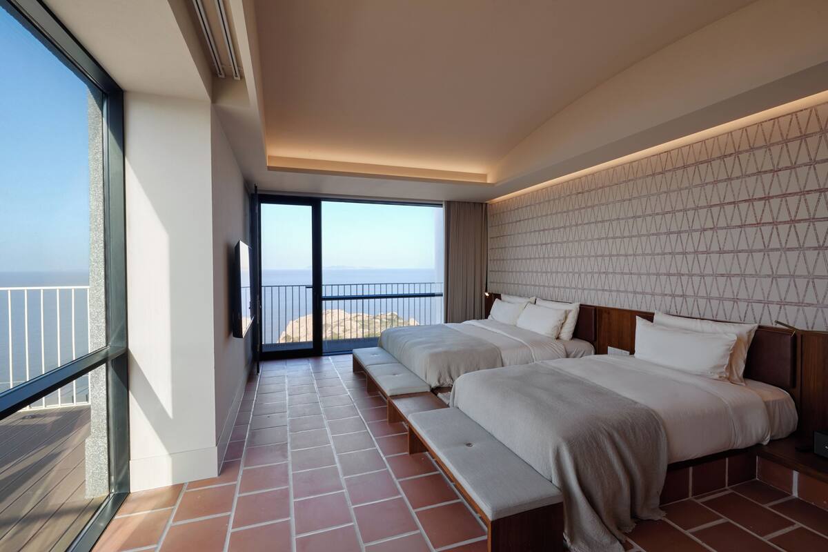 Habitación de hotel con dos camas, un ventanal grande y un balcón con vista al mar.