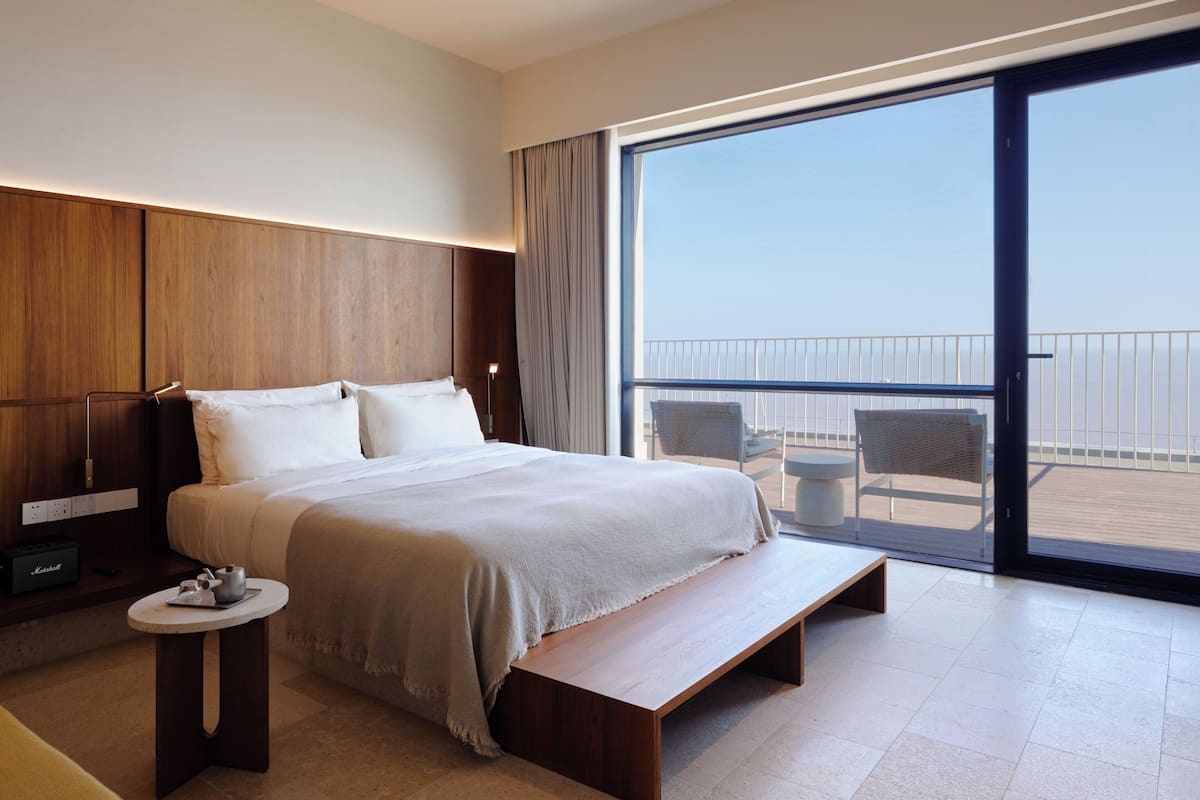Una habitación de hotel moderna con una cama grande, una mesita pequeña, un banco y un balcón con vistas.