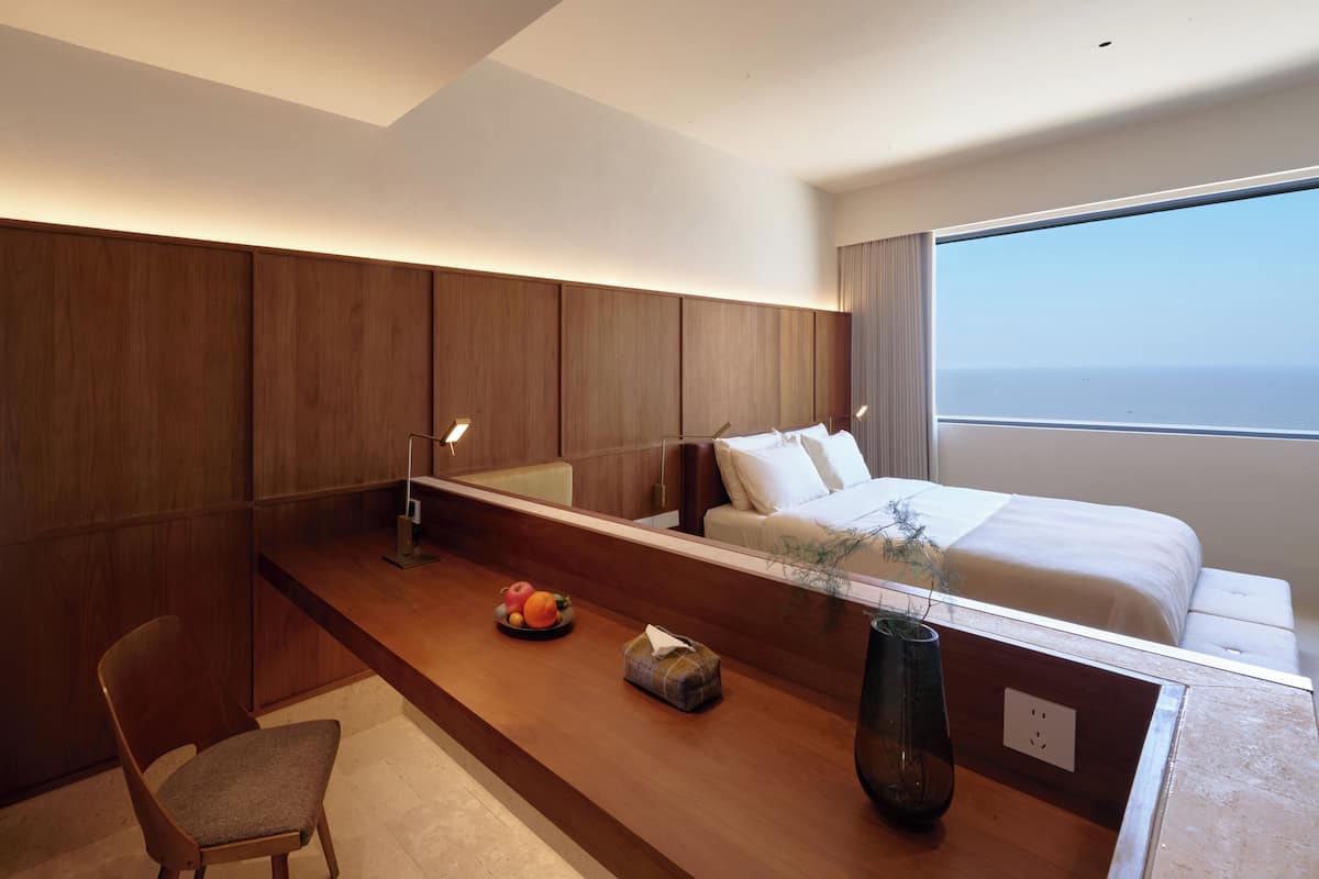 Una habitación de hotel moderna con un escritorio de madera, una silla, una mesita de noche con un florero, una cama con ropa blanca y un ventanal con vistas.