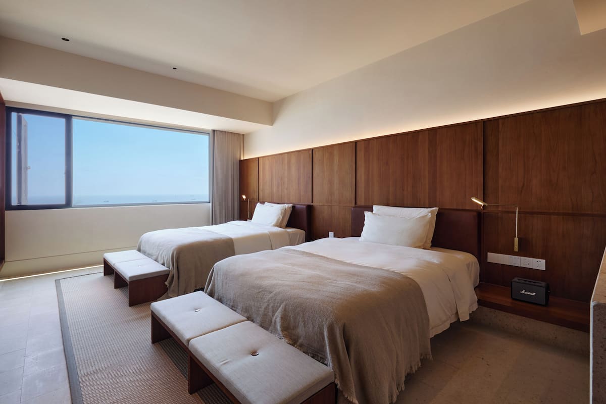 Una habitación de hotel moderna con dos camas, un ventanal grande, un cabecero de madera y un escritorio pequeño con una lámpara.