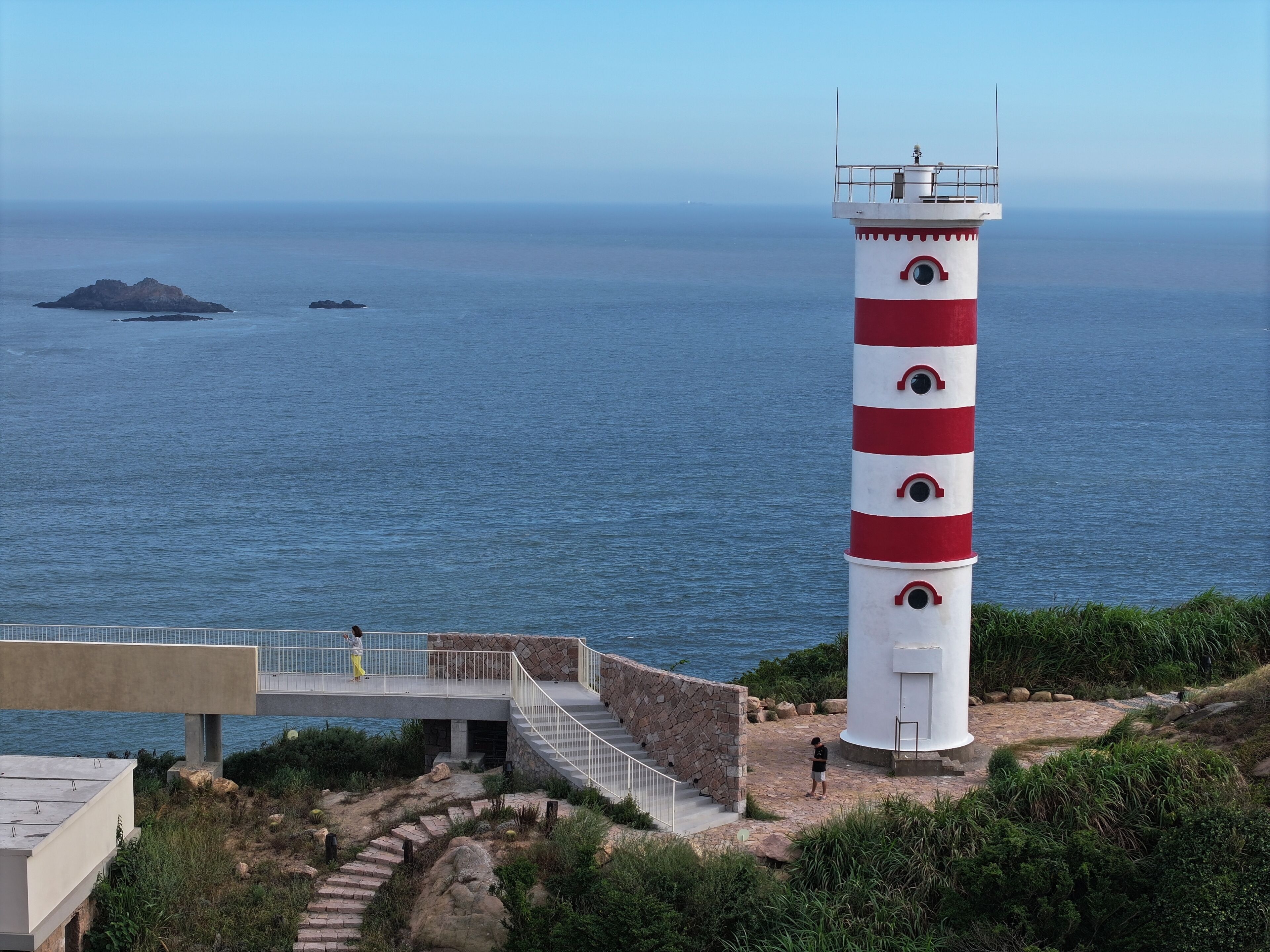 Un faro con exterior a rayas blancas y rojas, una escalera que conduce hasta él, y vista al mar con dos islas pequeñas a lo lejos.