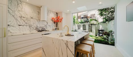 A modern kitchen with a marble backsplash, a central island with stools, and a view of a garden through a large window.