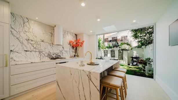 A modern kitchen with a marble backsplash, a central island with stools, and a view of a garden through a large window.