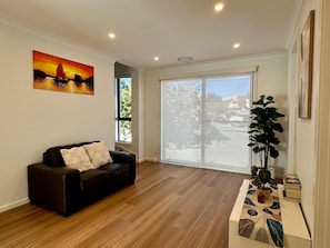 A living room with a black leather sofa, a painting of a sunset, a large window, and a potted plant. - Designer 4-Bed Coastal Home Walk To Beach & Shops (Maroubra)