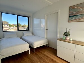 A bedroom with two single beds, a wooden dresser, a window with a view of buildings, and a door. - Designer 4-Bed Coastal Home Walk To Beach & Shops (Maroubra)