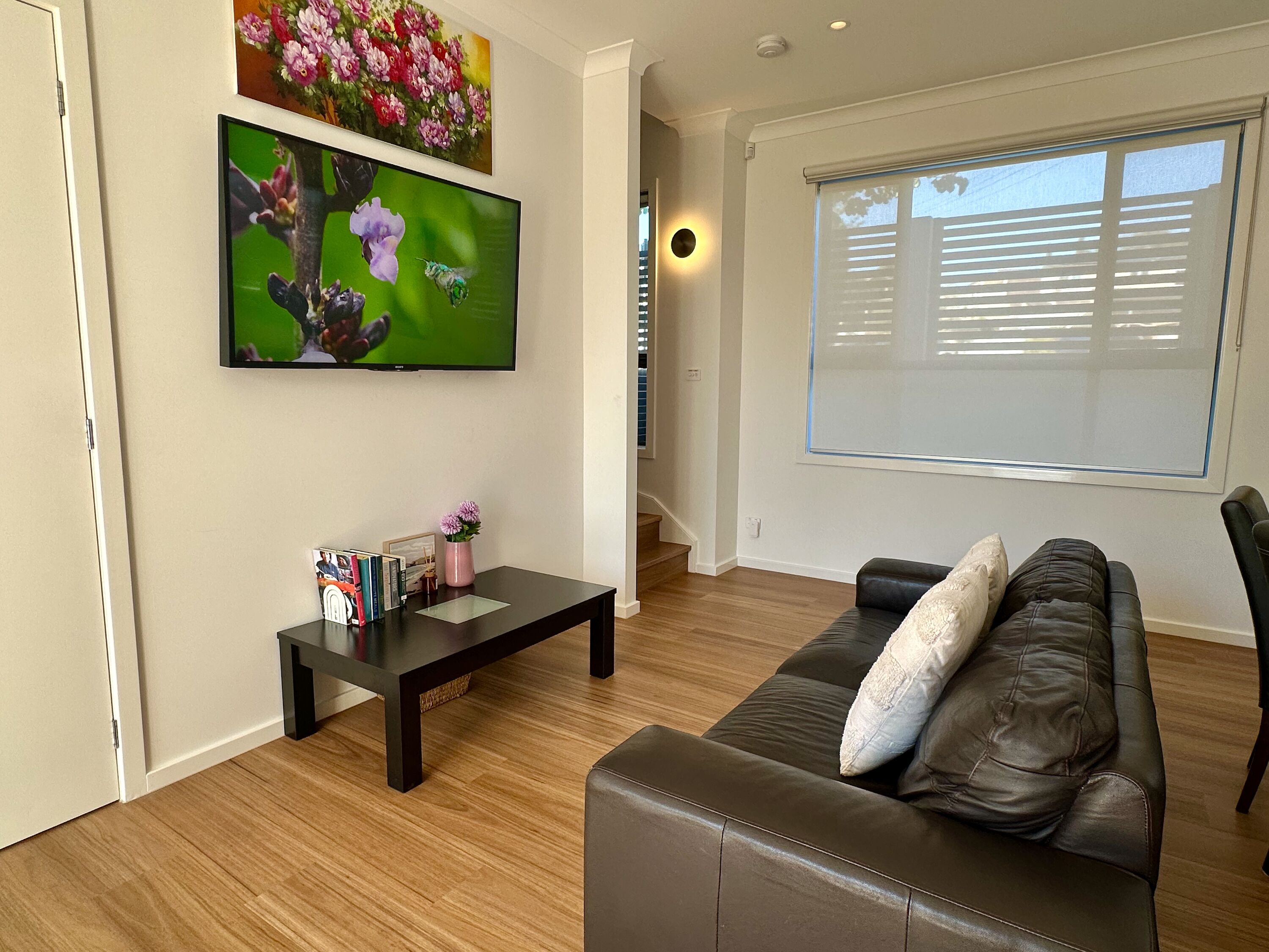 Una sala de estar con un sofá de cuero oscuro, una mesa de centro con libros y un florero, un televisor de pantalla plana en la pared y una ventana con persianas.