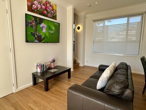 Een woonkamer met een donker leren bank, een bijzettafel met boeken en een vaas, een flatscreen televisie aan de muur en een raam met jaloezieën.