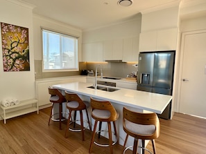 A modern kitchen with a central island, bar stools, stainless steel appliances, and a window. - Designer 4-Bed Coastal Home Walk To Beach & Shops (Maroubra)