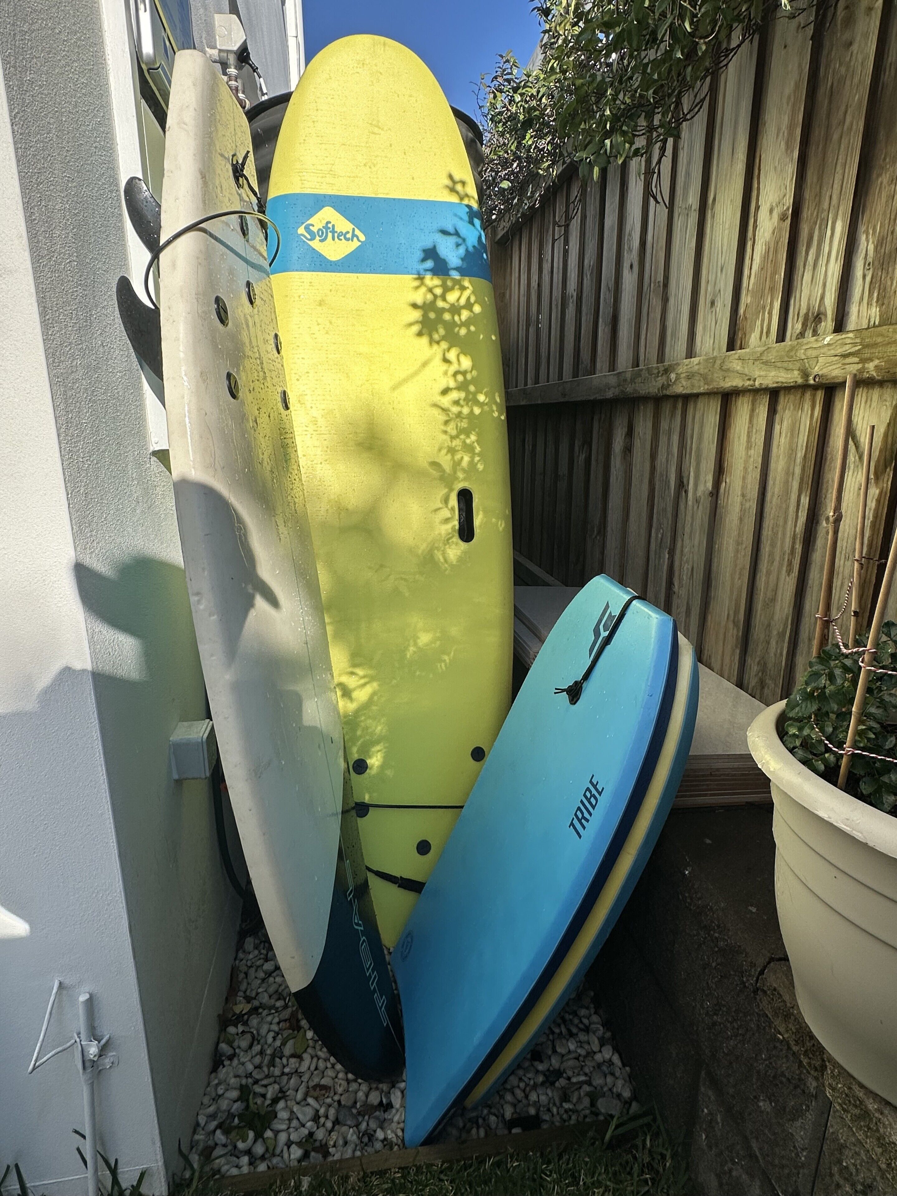 A surfboard leaning against a wall, a blue surfboard on the ground, and a wooden fence in the background.