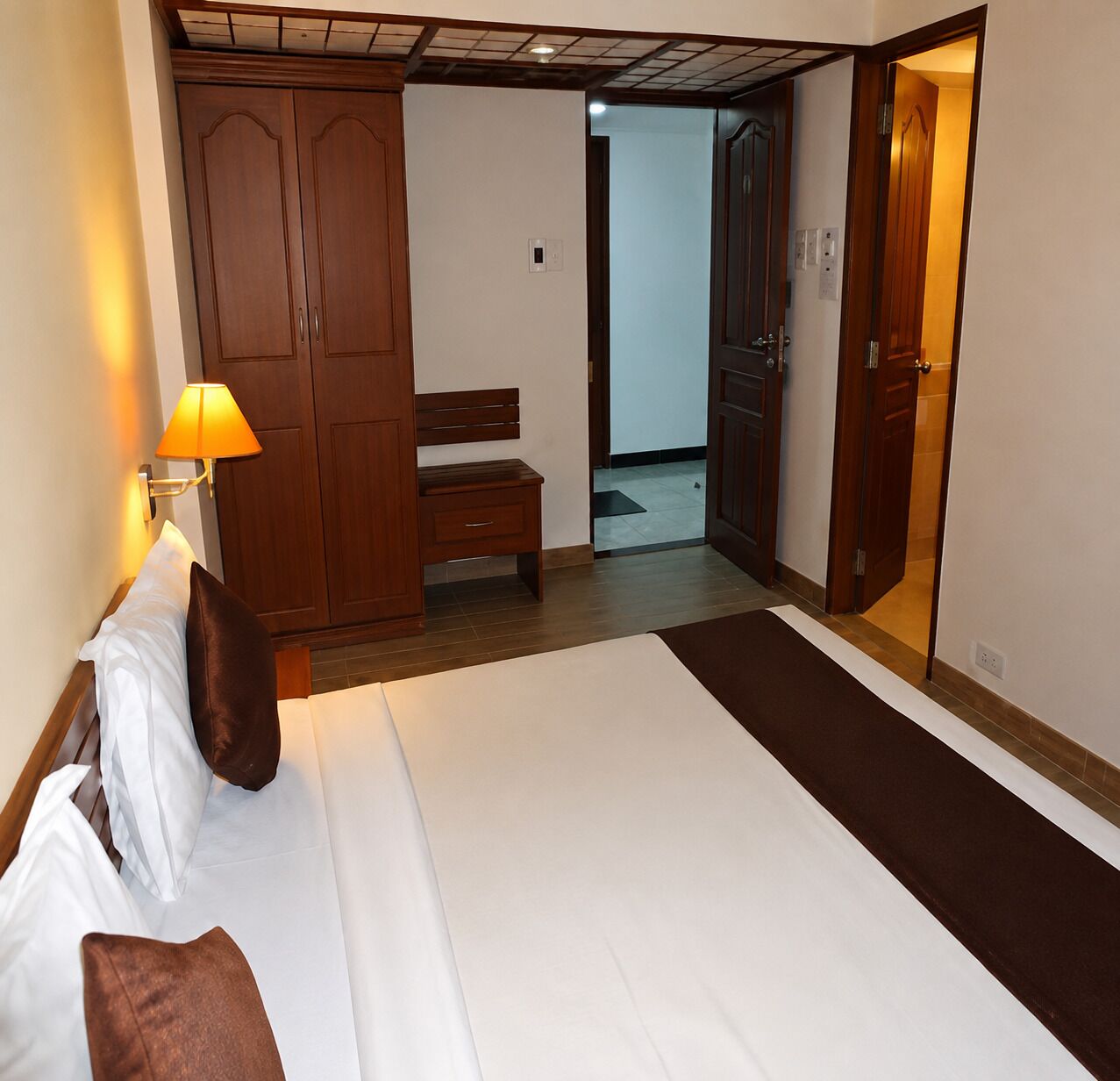 Habitación de hotel con cama, mesita de noche, lámpara, armario y un banco.