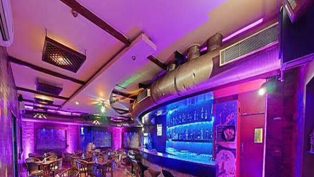 A bar area with wooden tables and chairs, a bar counter, and a large screen displaying a menu. - Hotel Royal Plazha (Chennai)
