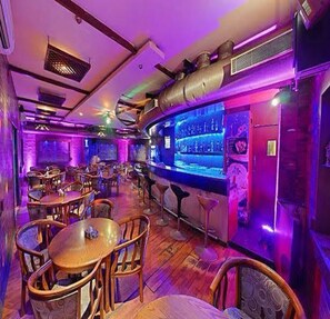 A bar area with wooden tables and chairs, a bar counter, and a large screen displaying a menu. - Hotel Royal Plazha (Chennai)