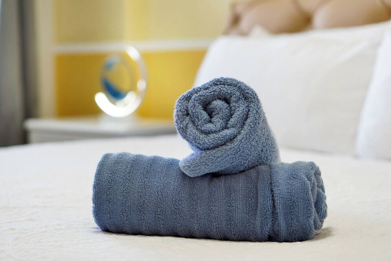 A neatly rolled towel on a bed with a white pillow and a round mirror in the background.