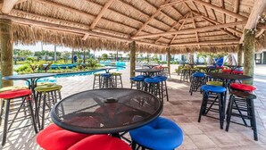 A covered outdoor seating area with tables and chairs, a pool in the background, and a thatched roof structure.