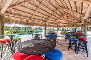 A covered outdoor seating area with tables and chairs, a pool in the background, and a thatched roof structure. - Themed Disney Villa 6BD Resort WaterSlide Pool Spa (Kissimmee)