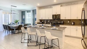 A modern kitchen with white cabinets, a central island with bar stools, stainless steel appliances, and a dining area with a table and chairs.