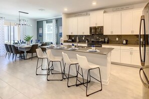 A modern kitchen with white cabinets, a central island with bar stools, stainless steel appliances, and a dining area with a table and chairs. - Themed Disney Villa 6BD Resort WaterSlide Pool Spa (Kissimmee)