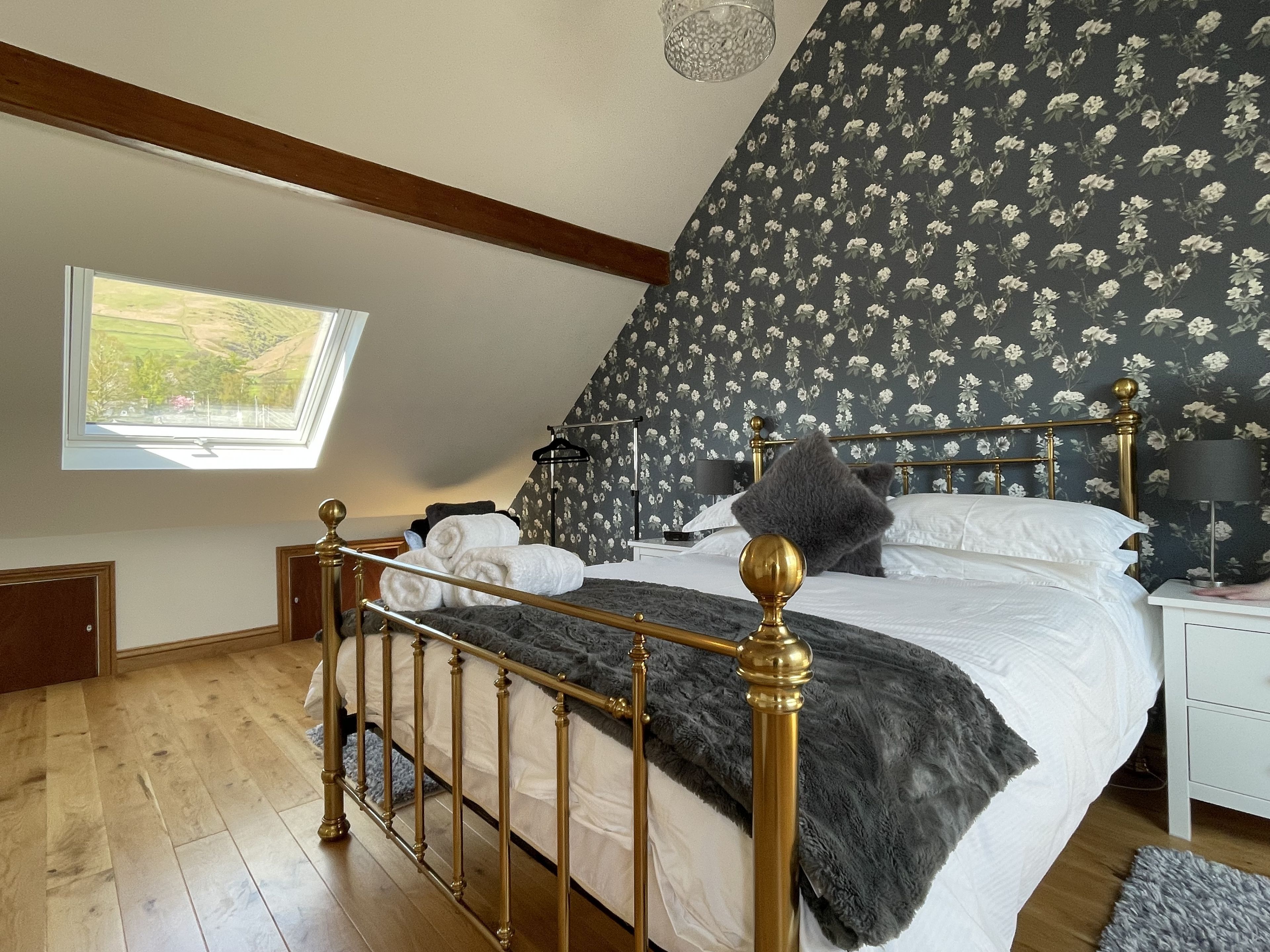 A bedroom with a sloped ceiling, a bed with a gold frame, a window with a view, and a floral wallpaper.
