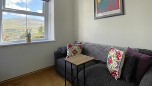 A room with a grey sofa, a small wooden table, a window with a view of hills, and a framed picture on the wall. - Ruby's Cottage -  a house that sleeps 4 guests  in 2 bedrooms (Threlkeld)