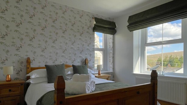 A bedroom with a wooden bed, floral wallpaper, a window with a view, and a bedside table with a lamp. - Ruby's Cottage -  a house that sleeps 4 guests  in 2 bedrooms (Threlkeld)