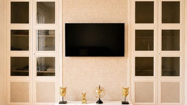 A modern living room with a flat-screen TV mounted on a brick wall, a white entertainment unit with decorative items, and a wooden floor. - Appartement 6 personnes  – Châtellerault (Châtellerault)