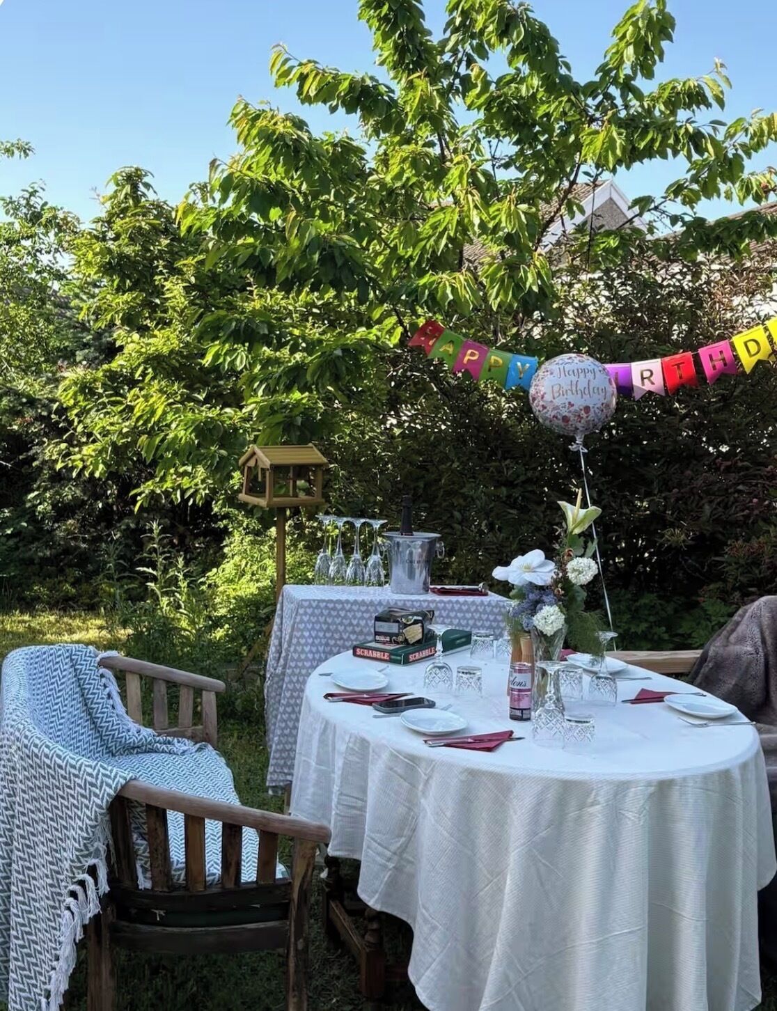 An outdoor dining area with round tables covered in white tablecloths, a birdhouse, and a 'Happy Birthday' banner.