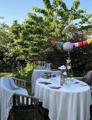 En udendørs spiseplads med runde borde dækket af hvide dugge, et fuglehus og et banner med teksten 'Happy Birthday'.