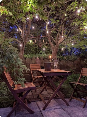 An outdoor seating area with a table and chairs under a tree, surrounded by stone walls and string lights.