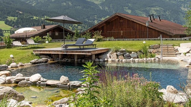 A pool area with lounge chairs, a wooden building, and a mountain backdrop. - 3A mit Tradition/am Bach by Interhome (Bad Hofgastein)